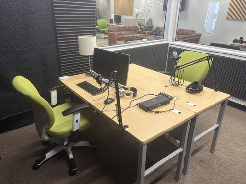 Podcast recording studio setup featuring a desk with a computer, microphones, and green ergonomic chairs.