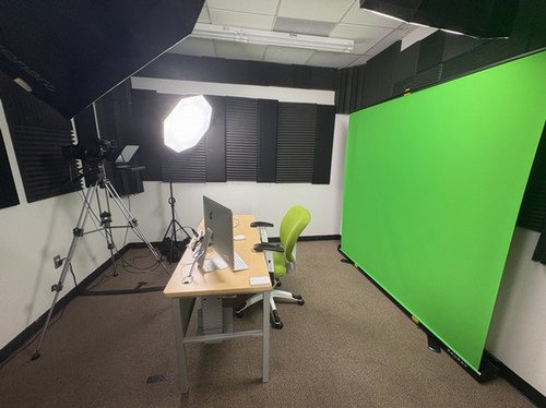 A media production workspace featuring a green screen, professional lighting, and a desk with a computer and chair.